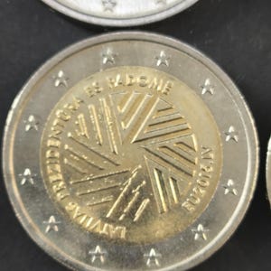 Può includere: Primo piano di una moneta da due euro con un disegno dorato al centro e un anello esterno argentato. La moneta presenta testo in lettone e l'anno 2013. Fa parte di una collezione di monete.