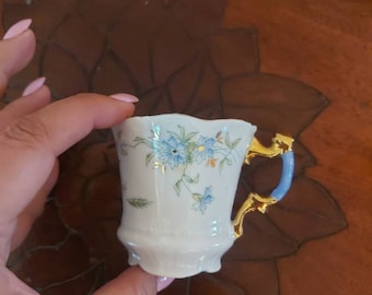 Könnte beinhalten: Eine kleine weiße Porzellantasse mit einem blauen und goldenen Blumendesign. Die Tasse hat einen goldenen Rand und einen blauen Henkel.