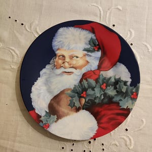 May include: A blue and white ceramic plate featuring a festive image of Santa Claus holding a sprig of holly. The plate has a red hat, white beard, and a blue background.