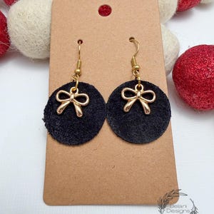 May include: A pair of black suede earrings with gold-colored bow charms. The earrings are circular and hang from gold-colored hooks. The earrings are displayed on a brown card.