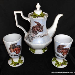 Può includere: Un servizio da tè in ceramica bianca composto da una teiera e due tazze. La teiera e le tazze sono decorate con un disegno di scoiattolo marrone su uno sfondo innevato. Le basi delle tazze e della teiera presentano un motivo a foglie verdi e nere.