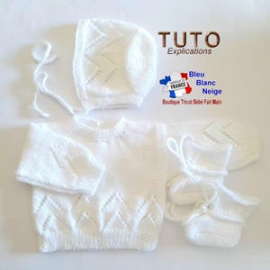 Peut inclure: Un ensemble de vêtements pour bébé en tricot blanc, comprenant un pull, un bonnet, des moufles et des chaussons. L'ensemble présente un motif en losange et des liens. L'image comprend également le texte "TUTO Explications" et un logo du drapeau français.