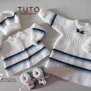 Peut inclure: Un ensemble bébé en tricot blanc avec des rayures bleues et blanches. L'ensemble comprend un bonnet, un cardigan, une robe et des chaussons.