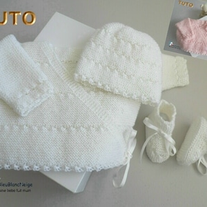 May include: A white knitted baby outfit set with a hat, sweater, and booties. The set is made with a delicate stitch pattern and features a white satin ribbon.