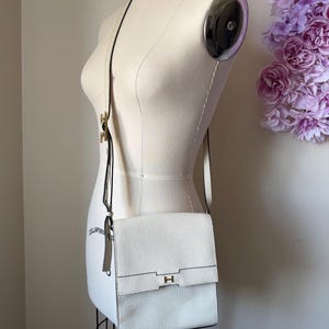 May include: A white leather crossbody bag with a textured finish, featuring a rectangular flap closure secured by a gold-toned metal H-shaped clasp. The bag includes a thin, adjustable shoulder strap and a matching leather tag attached to the side.
