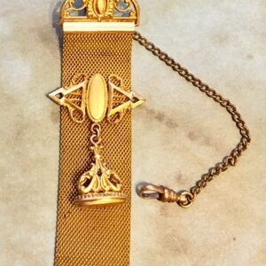 May include: A gold chain with a decorative fob. The fob is made of a mesh material and has a gold pendant with a crown design. The chain is attached to a swivel clasp.
