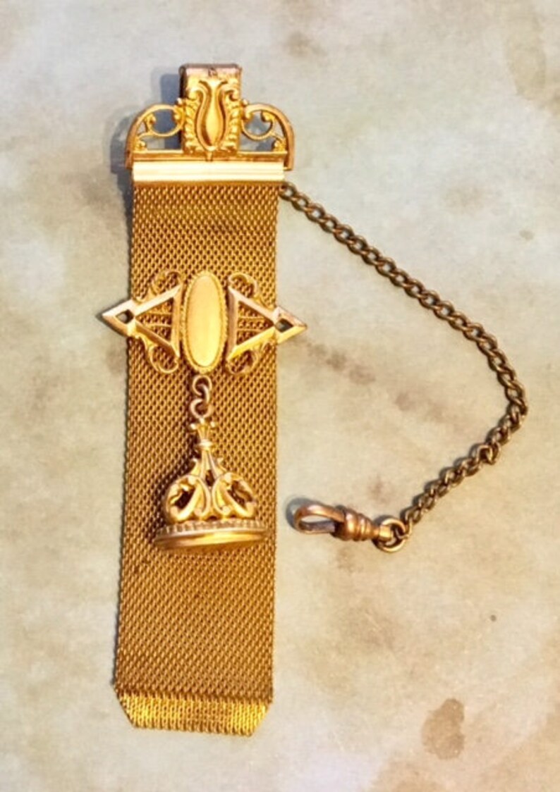 May include: A gold chain with a decorative fob. The fob is made of a mesh material and has a gold pendant with a crown design. The chain is attached to a swivel clasp.