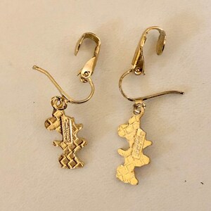 May include: A pair of gold-coloured earrings with an unusual, abstract design. Each earring has a hook closure and a dangling charm with a textured, geometric pattern. The charms have a small, rectangular marking.