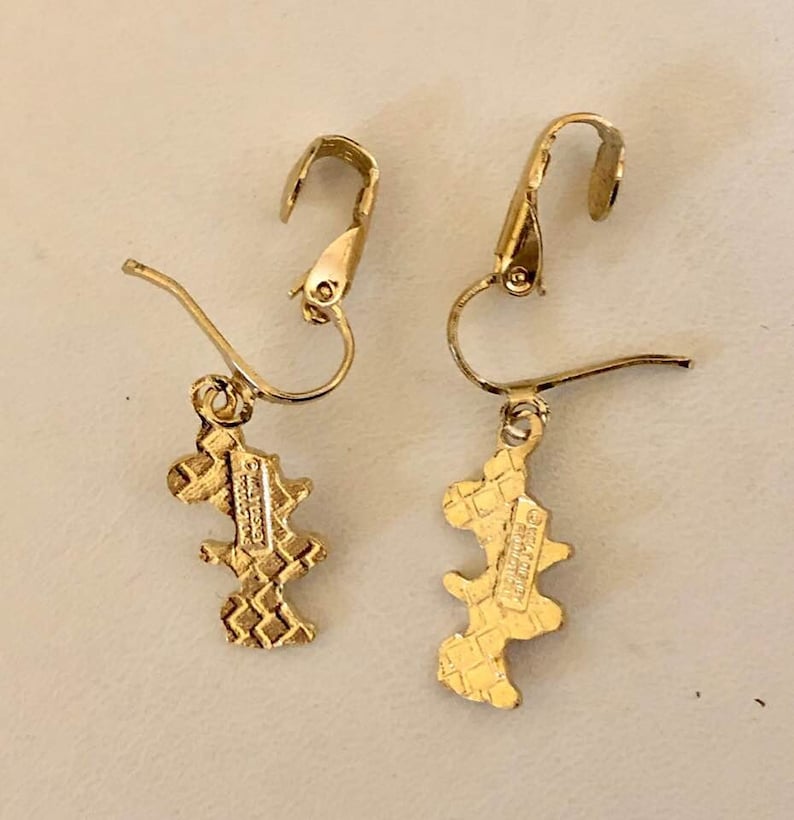 May include: A pair of gold-coloured earrings with an unusual, abstract design. Each earring has a hook closure and a dangling charm with a textured, geometric pattern. The charms have a small, rectangular marking.