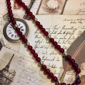 May include: A red glass bead necklace with gold accents. The necklace is lying on a table with vintage postcards and a clock.