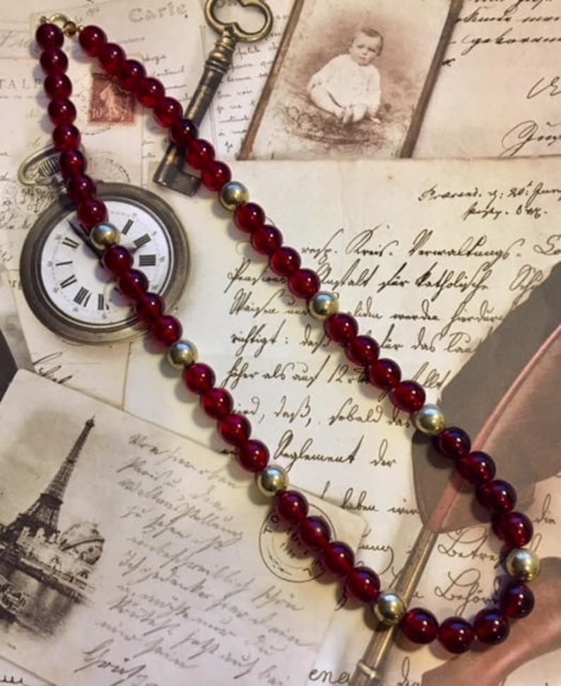 May include: A red glass bead necklace with gold accents. The necklace is lying on a table with vintage postcards and a clock.