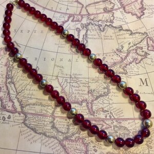 May include: A red and gold beaded necklace with a vintage feel. The necklace is made of round, red glass beads and gold metal beads. The necklace is lying on a map.