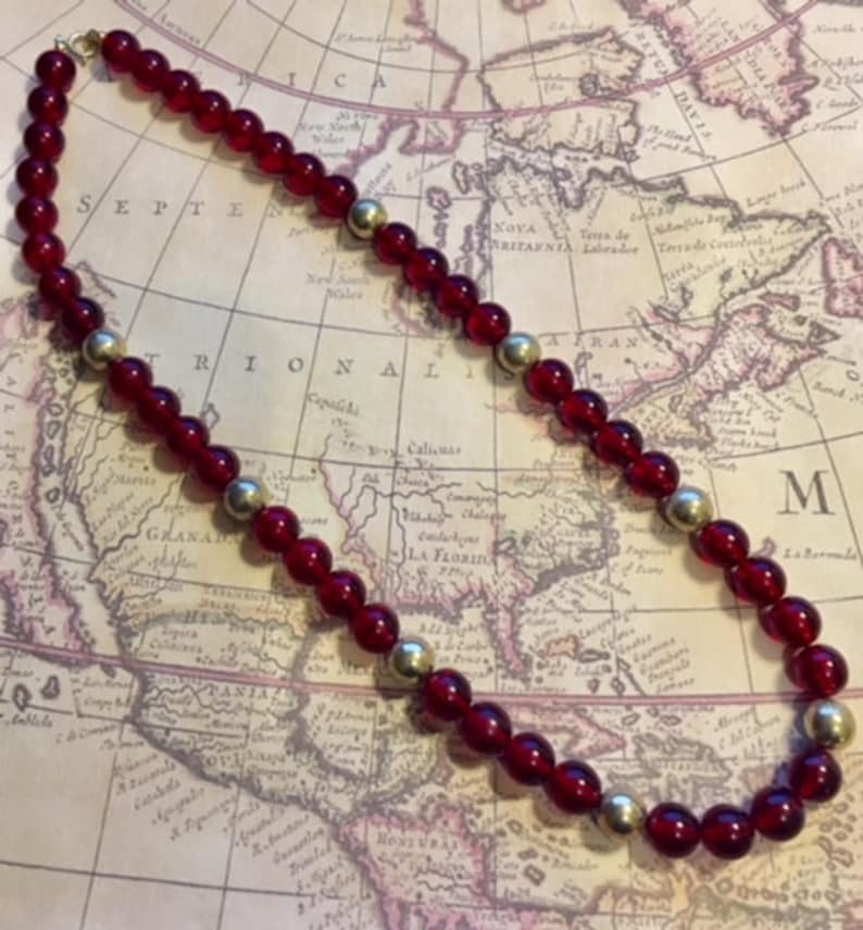 May include: A red and gold beaded necklace with a vintage feel. The necklace is made of round, red glass beads and gold metal beads. The necklace is lying on a map.