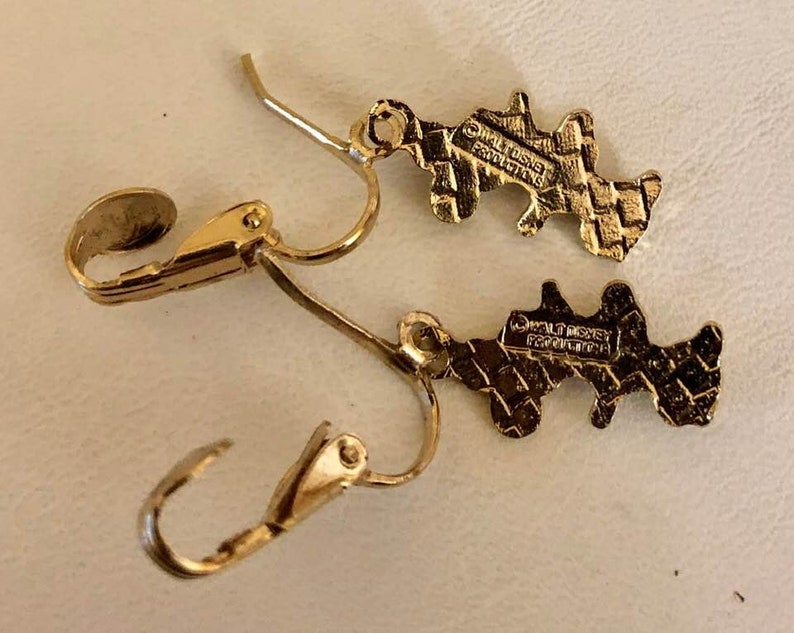May include: A pair of gold-coloured earrings with a distinctive shape. The earrings feature a lever-back clasp and a textured pattern. The reverse of each earring is stamped with "&copy; WALT DISNEY PRODUCTIONS".