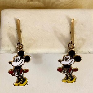 May include: A pair of gold-coloured earrings featuring a cartoon mouse character with black ears, a white face, red jacket, and yellow shoes. The earrings are hanging from a gold-coloured post.