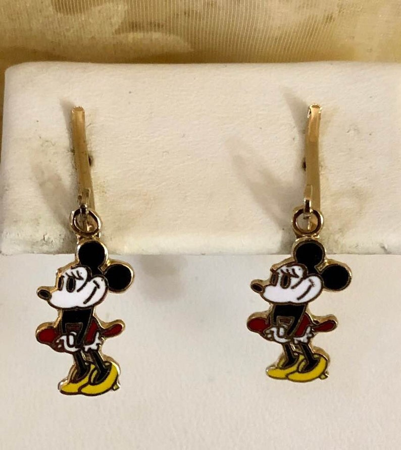 May include: A pair of gold-coloured earrings featuring a cartoon mouse character with black ears, a white face, red jacket, and yellow shoes. The earrings are hanging from a gold-coloured post.
