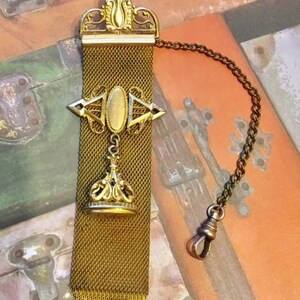 May include: A gold-toned pocket watch chain with a decorative fob. The chain is made of a fine mesh and has a decorative pendant with a crown design. The fob has a small oval-shaped piece of glass or metal in the center.