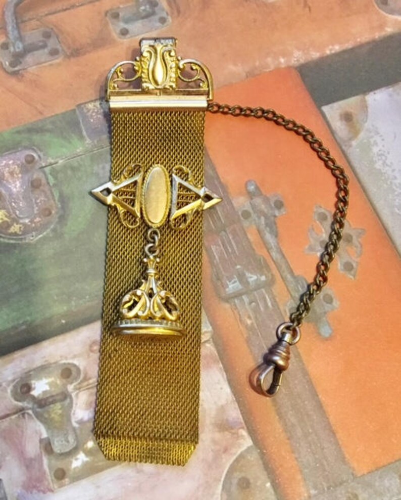 May include: A gold-toned pocket watch chain with a decorative fob. The chain is made of a fine mesh and has a decorative pendant with a crown design. The fob has a small oval-shaped piece of glass or metal in the center.