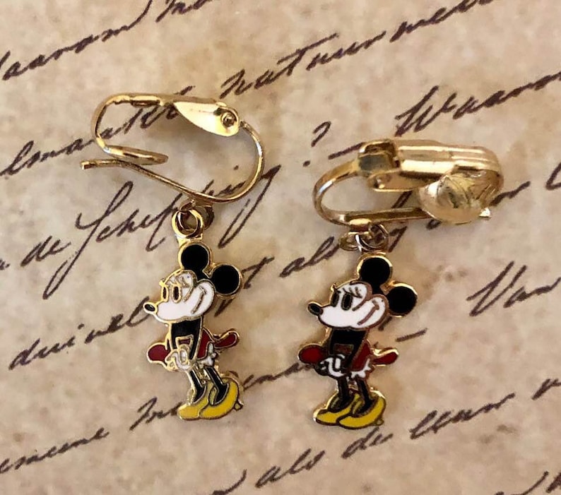 May include: Gold-coloured clip-on earrings with a Mickey Mouse design. Each earring has a black head, white face, red jacket, and yellow shoes. The earrings are set against a vintage paper background.