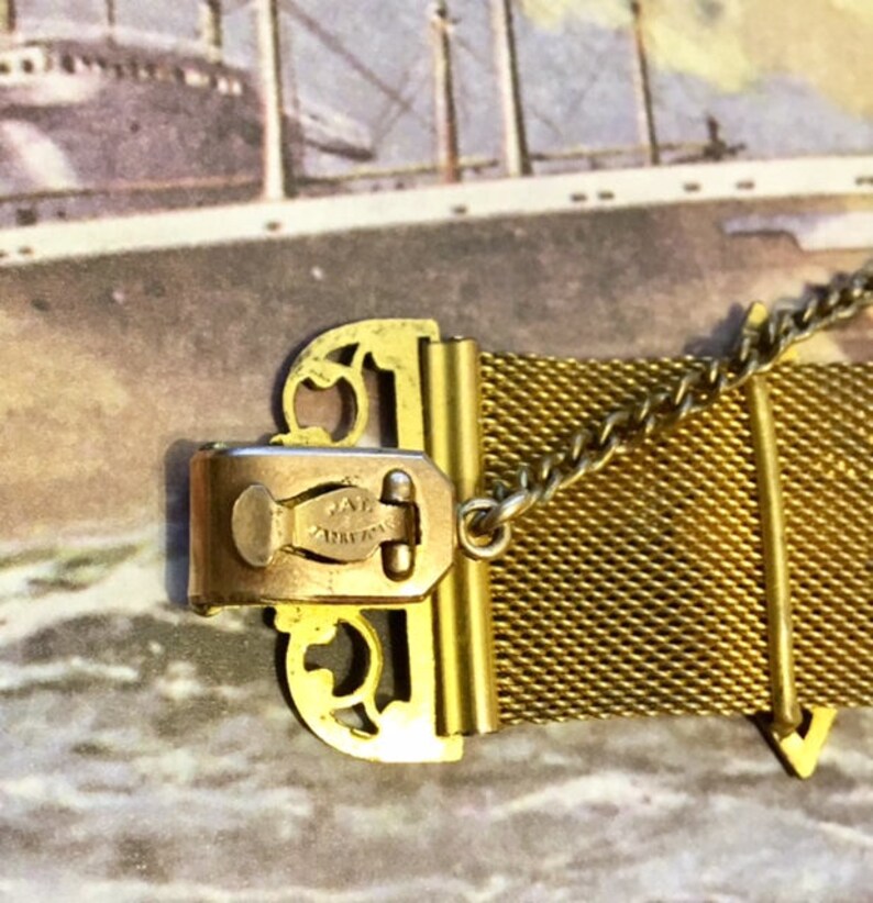 May include: Gold-toned metal mesh bracelet with a decorative clasp. The clasp has a filigree design and the words "Jae  Ancre" engraved on it.