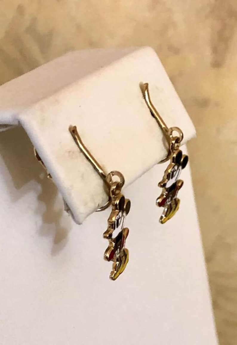 May include: Gold-coloured dangle earrings with a Mickey Mouse design. The earrings have a hook closure and feature Mickey's iconic face with black, white, red, and yellow enamel details. The earrings are displayed on a white surface.