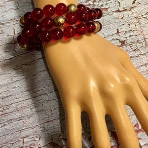 May include: A multi-strand bracelet with red glass beads and gold accents. The bracelet is worn on a mannequin hand.