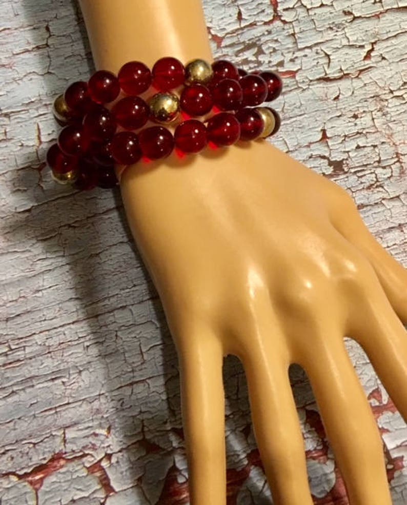 May include: A multi-strand bracelet with red glass beads and gold accents. The bracelet is worn on a mannequin hand.