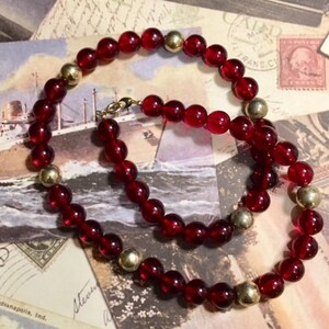 May include: A red glass bead necklace with gold accents. The necklace is made of round beads and is strung on a gold chain.