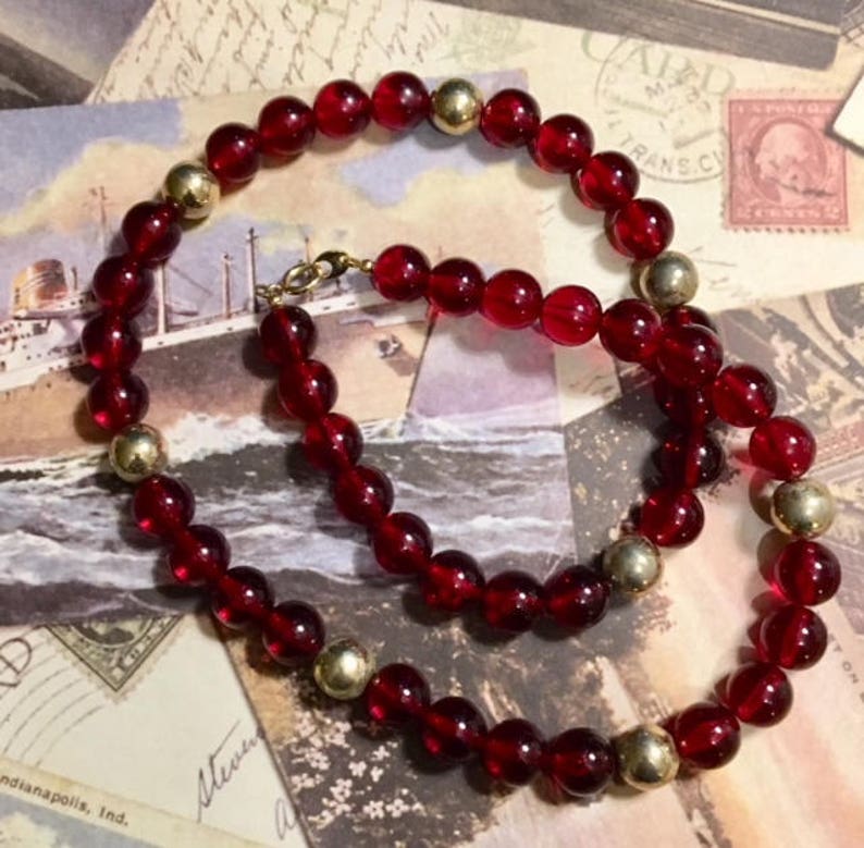 May include: A red glass bead necklace with gold accents. The necklace is made of round beads and is strung on a gold chain.