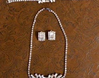 Op de afbeelding: Een vintage set met strass sieraden met een ketting, armband en oorbellen. De set heeft een geometrisch ontwerp met heldere strass.