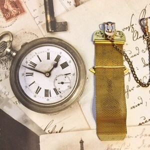 May include: A gold pocket watch chain with a decorative clasp and a mesh chain. The chain is attached to a pocket watch with a white face and black Roman numeral hour markers. The watch is lying on a piece of paper with writing on it.