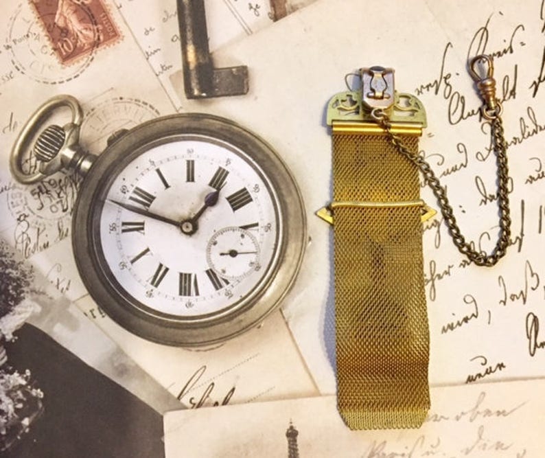 May include: A gold pocket watch chain with a decorative clasp and a mesh chain. The chain is attached to a pocket watch with a white face and black Roman numeral hour markers. The watch is lying on a piece of paper with writing on it.