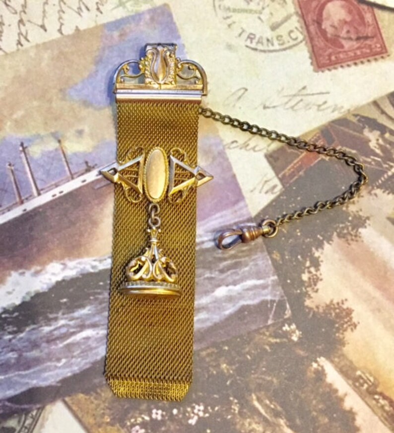 May include: A gold chain with a decorative fob. The fob is made of a mesh material and has a gold crown-shaped pendant. The chain is attached to a clasp.