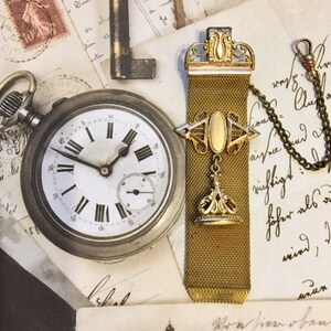 May include: A gold chain watch fob with a decorative top and a chain attached. The fob has a mesh design and a small, round, gold-colored pendant at the bottom. The fob is lying on a piece of paper with writing on it.