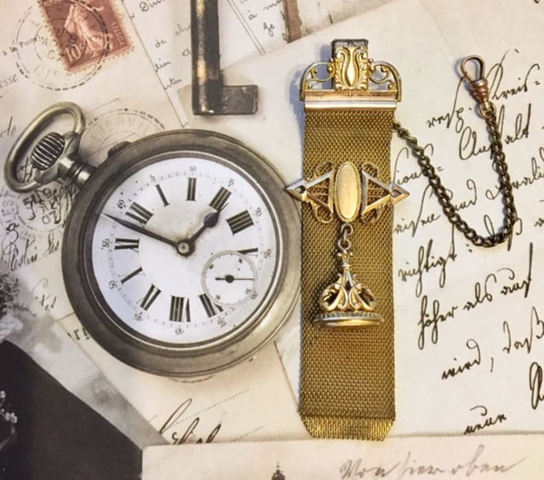 May include: A gold chain watch fob with a decorative top and a chain attached. The fob has a mesh design and a small, round, gold-colored pendant at the bottom. The fob is lying on a piece of paper with writing on it.