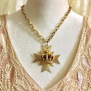 May include: A gold chain necklace with a large pendant featuring a gold crown with red jewels on top of a gold cross.