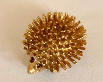May include: A gold-toned brooch in the shape of a hedgehog. The hedgehog has a spiky body and a small face with two orange rhinestones for eyes.