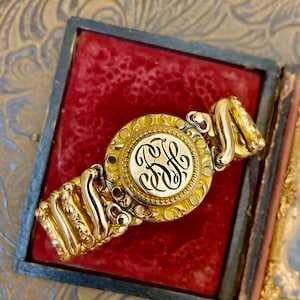 May include: A gold-toned antique bracelet with an engraved monogram in a round, ornate setting. The bracelet is displayed in a velvet-lined box with a red interior. The design features intricate detailing and a classic aesthetic.