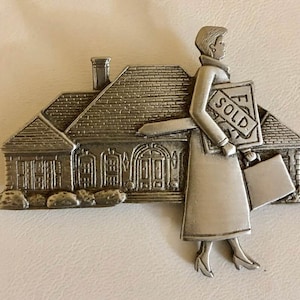 May include: A silver-toned metal figurine depicting a house with a woman holding a "SOLD" sign and a briefcase. The house has detailed architectural features, including a chimney and multiple windows. The woman is wearing a coat and heels.