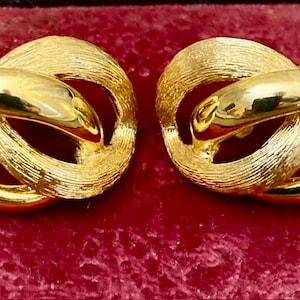 Op de afbeelding: Een paar gouden oorbellen met een geknoopt ontwerp. De oorbellen zijn gemaakt van metaal en hebben een geborstelde afwerking.