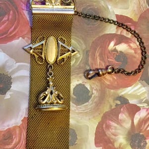 May include: A gold-toned chain with a mesh fob and a decorative pendant. The pendant features a crown-like design with a gold oval in the center.