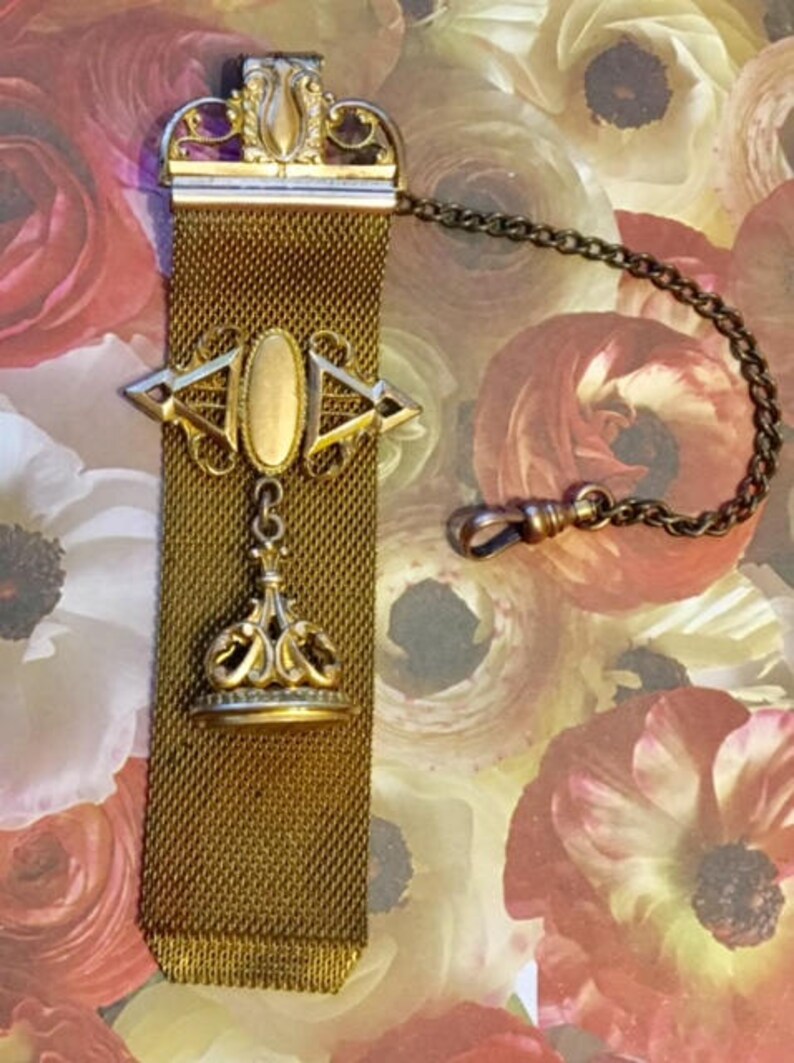 May include: A gold-toned chain with a mesh fob and a decorative pendant. The pendant features a crown-like design with a gold oval in the center.