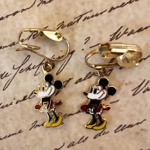 May include: Gold-coloured clip-on earrings with a Mickey Mouse design. Each earring has a black head, white face, red shorts, and yellow shoes. The earrings are set against a vintage paper background.