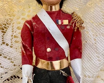 Könnte beinhalten: Eine Puppe, die als britischer Soldat verkleidet ist, trägt eine rote Jacke mit goldenen Verzierungen, einen schwarzen Pelz Hut mit einer weißen Feder und eine schwarze Hose. Die Puppe hält eine silberne Trompete.