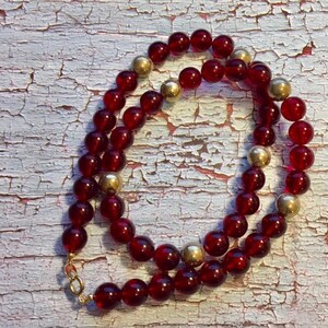 May include: A red glass bead necklace with gold accents. The necklace is made of round beads and has a gold clasp.