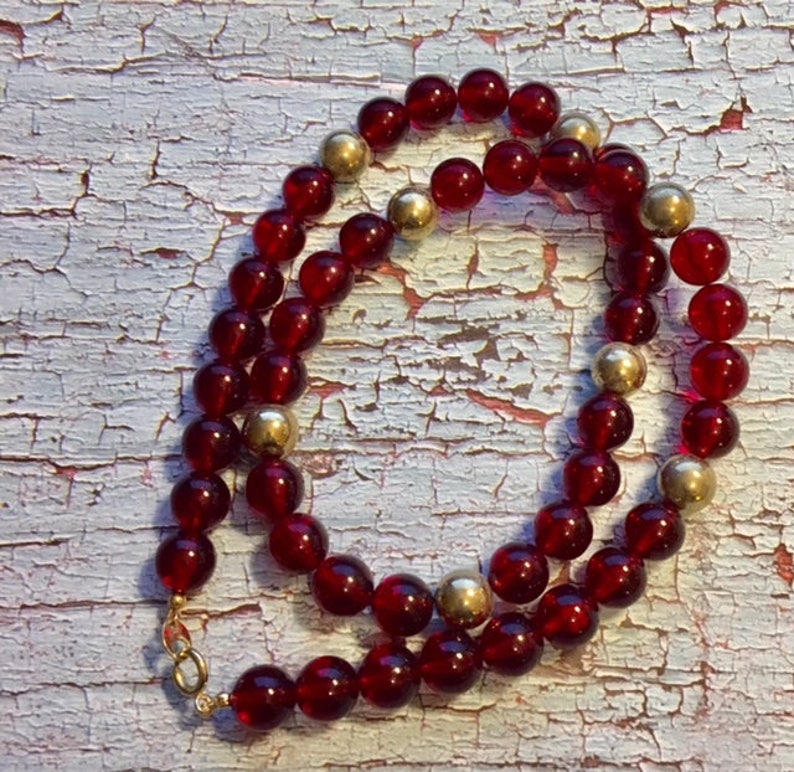 May include: A red glass bead necklace with gold accents. The necklace is made of round beads and has a gold clasp.