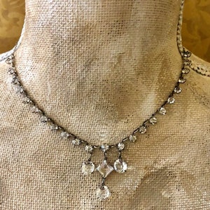 May include: A silver necklace with a dangling crystal pendant. The pendant features a square-shaped crystal in the centre, surrounded by smaller round crystals.