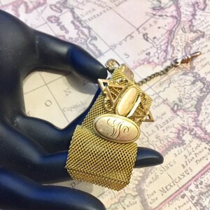 May include: A gold-toned chain with a mesh fabric fob and two oval-shaped charms. The charms have a gold-toned filigree design and one has an engraved letter 'S'.