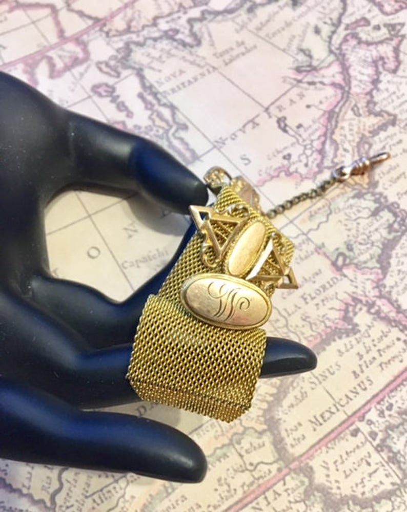 May include: A gold-toned chain with a mesh fabric fob and two oval-shaped charms. The charms have a gold-toned filigree design and one has an engraved letter 'S'.