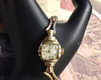 May include: A gold-toned wristwatch with a white face and black Roman numeral hour markers. The watch has a gold-toned metal band with a decorative design.